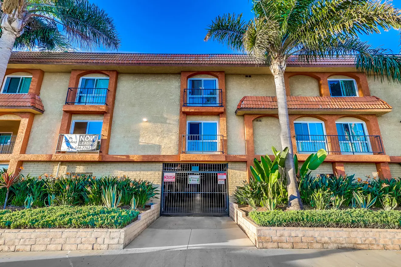 A beige two-story building with orange trim, balconies, and a Now Leasing sign. Palm trees and lush greenery frame the entrance, conveying a sunny, inviting atmosphere.