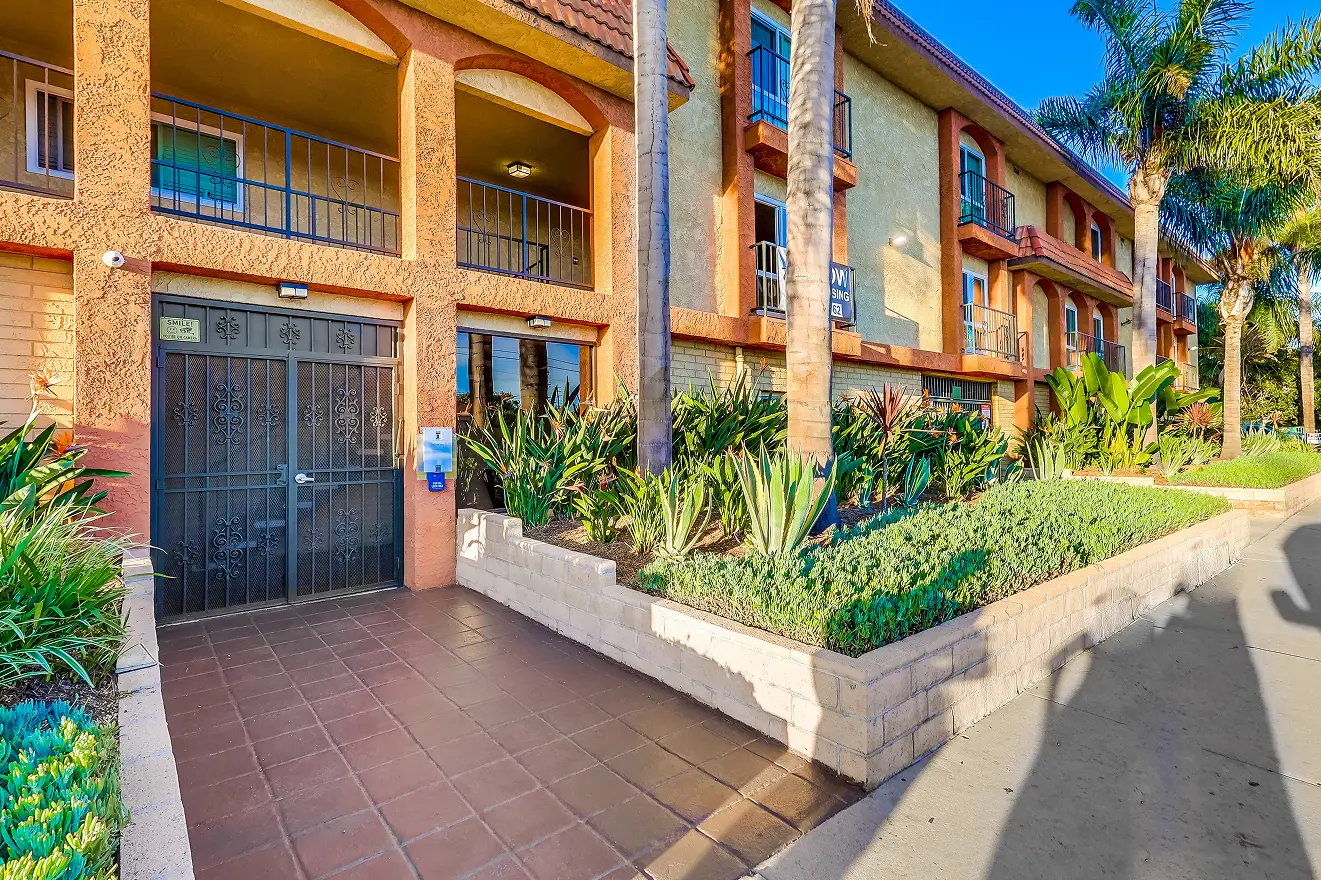 Apartment building entrance with arched stone facade, metal gate, and neatly landscaped greenery. Sunlit path creates a welcoming, inviting atmosphere.