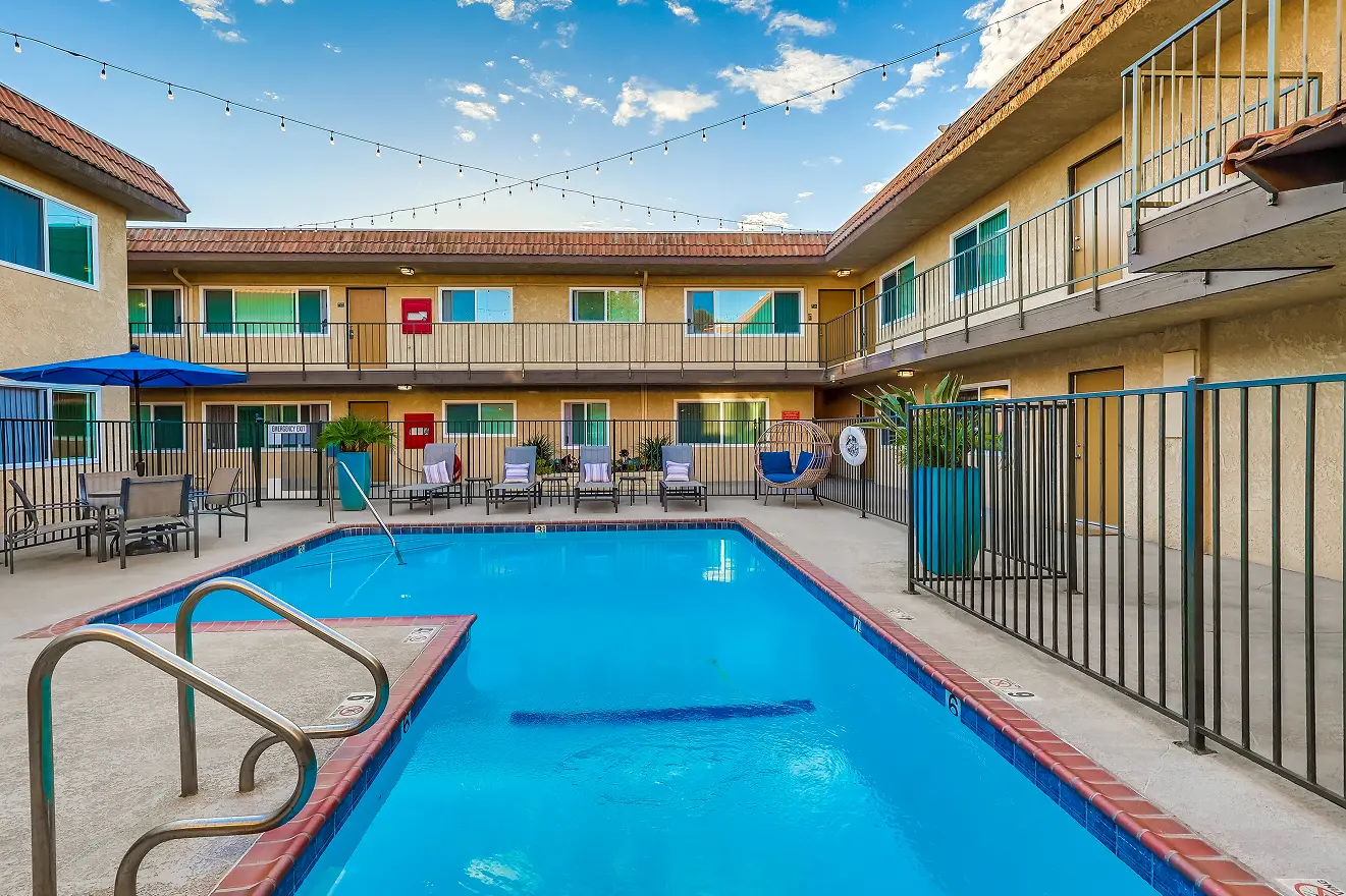 A courtyard pool surrounded by a two-story motel, with lounge chairs and umbrellas. String lights add a relaxed and inviting atmosphere under a blue sky.
