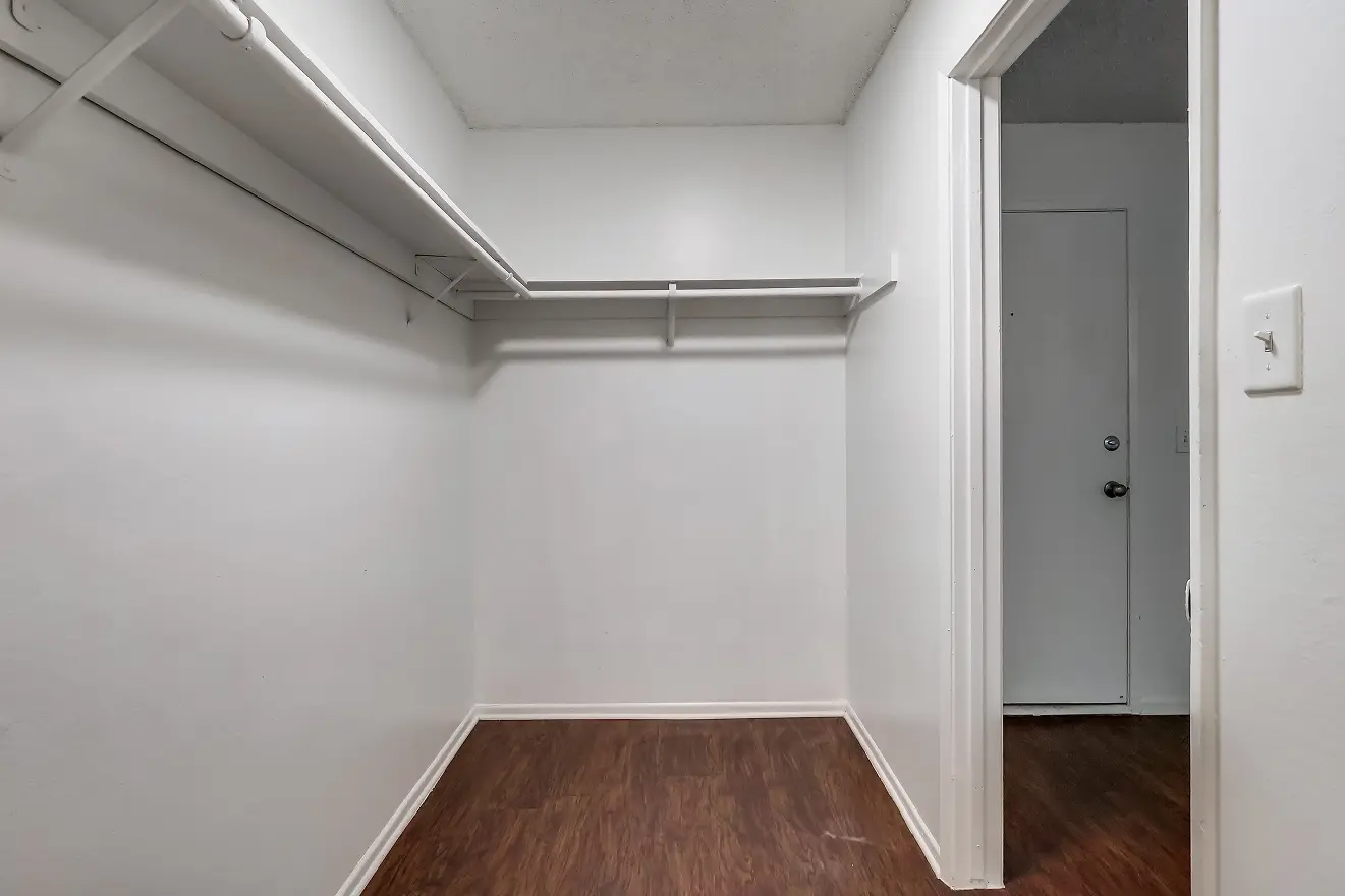 Empty walk-in closet with white walls, wooden floor, and shelves along the left and back wall, near an open door in a residential space.